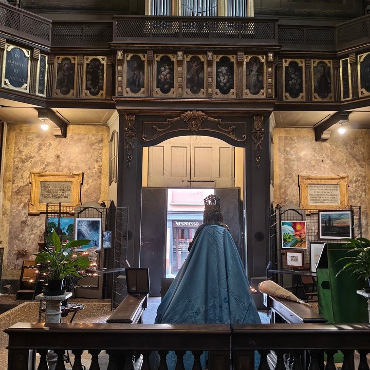 Interno di chiesa: la statua della Madonna con il manto azzurro, di spalle, guarda il portone aperto sulla strada con l'insegna Nespresso