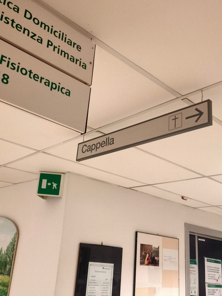 Corridoio d'ospedale: il cartello sospeso «Cappella» con la croce e la freccia, accanto all'uscita di emergenza