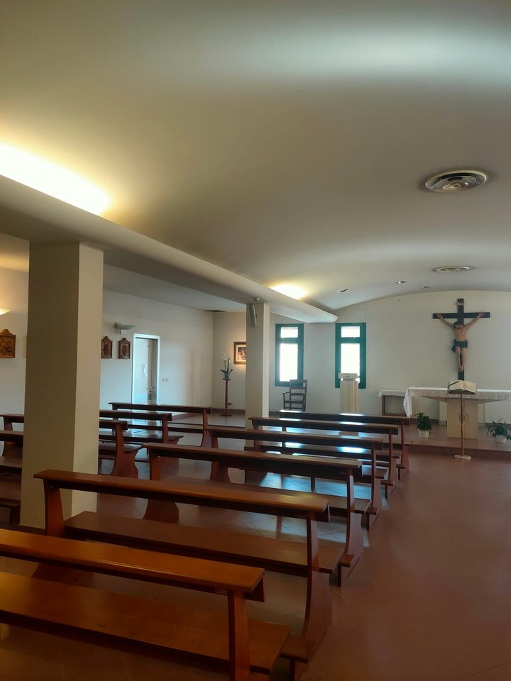 Interno della cappella dell'ospedale: banchi vuoti, crocifisso sopra l'altare, ambone e leggio